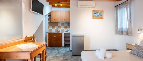 A bedroom with a bed, a desk, a chair, a television, a refrigerator, and a kitchen area in the background.
