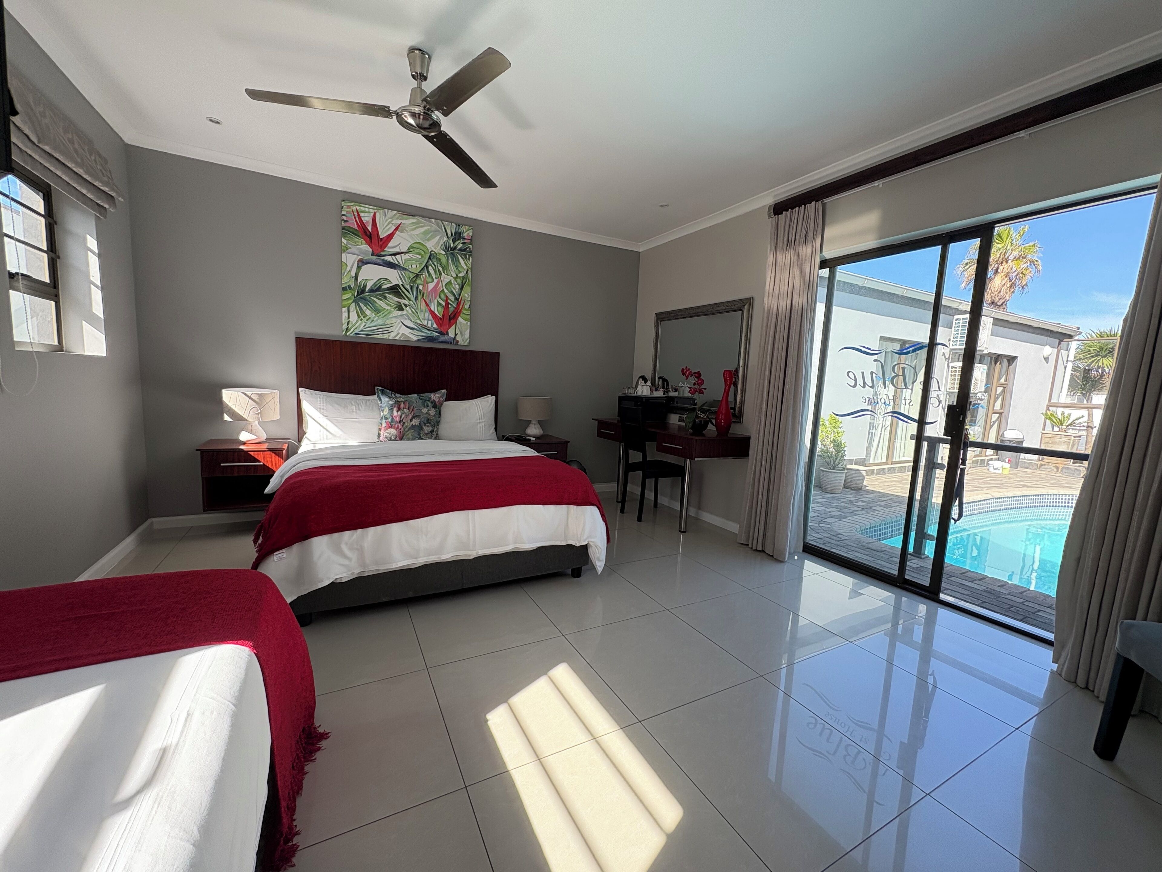 A hotel room with a bed, a desk, a chair, a desk lamp, a ceiling fan, a painting, and a view of a swimming pool.