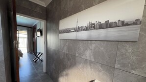 A hallway with grey tiled walls, a cityscape artwork, and a door leading to a room with a bed and a window. - Le Blue Guesthouse (Gqeberha (Port Elizabeth))