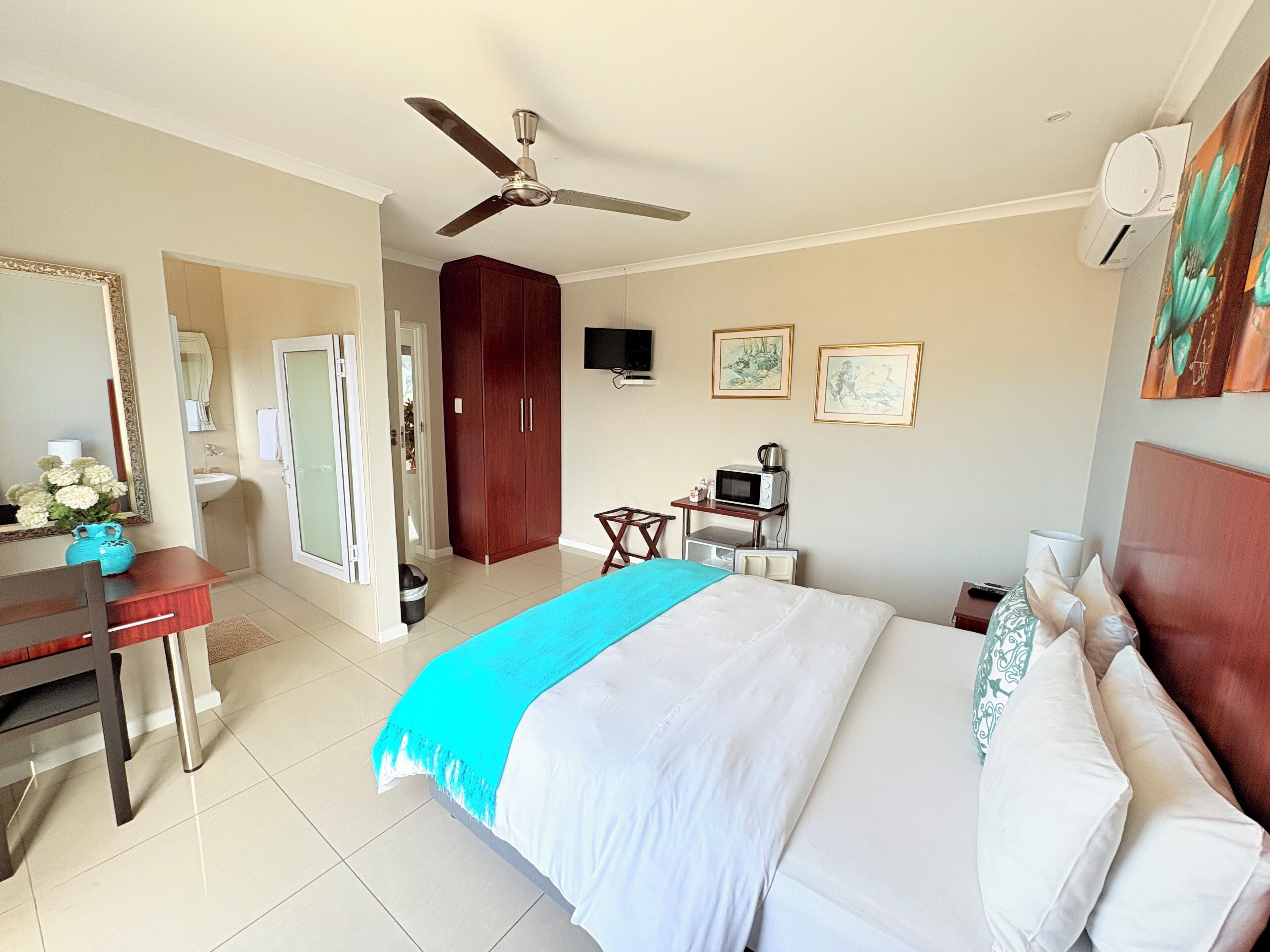 Habitación de hotel con cama, escritorio, armario, ventilador de techo, televisión y baño visible a través de una puerta abierta.