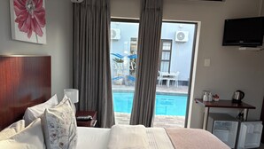 A hotel room with a bed, a desk, a TV, and a view of a swimming pool. - Le Blue Guesthouse (Gqeberha (Port Elizabeth))