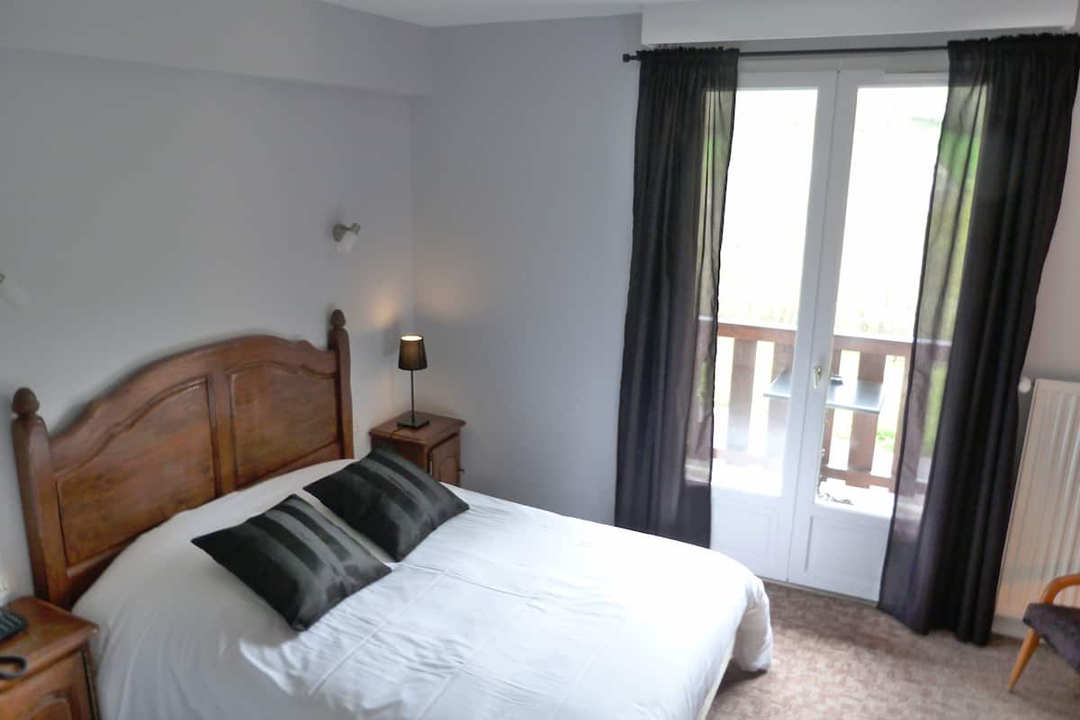 Ein Schlafzimmer mit einem hölzernen Kopfteil, einer weißen Bettdecke, zwei schwarz-weiß gestreiften Kissen, einer Nachttischlampe und Blick auf einen Balkon durch eine Glastür.