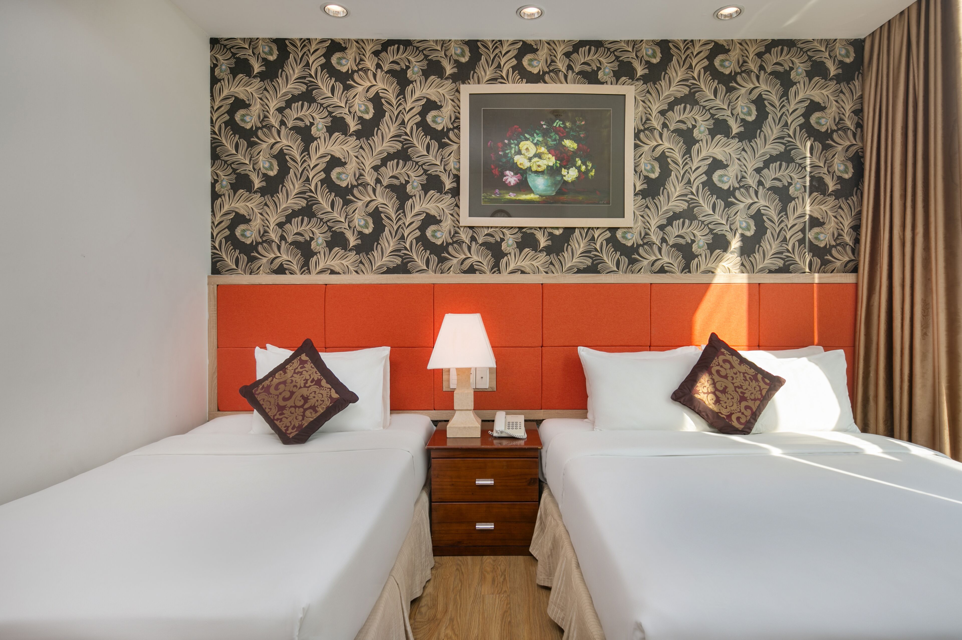 A hotel room with two beds, a nightstand with a lamp, a framed floral artwork on the wall, and a wooden floor.