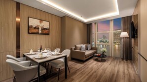 A modern hotel room with a dining area, a sofa, and a large window offering a city view. - Sun River Hotel (Da Nang)