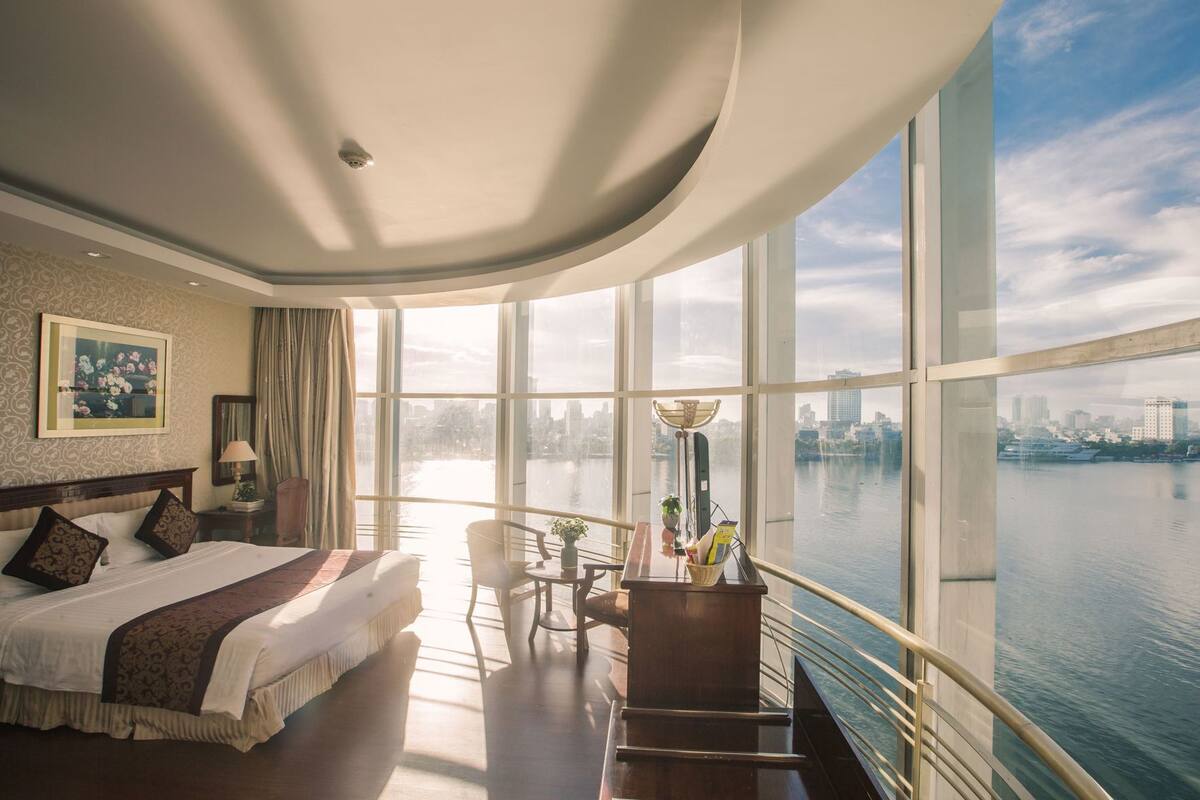 A hotel room with a large bed, a desk, a chair, and a view of a cityscape and water.