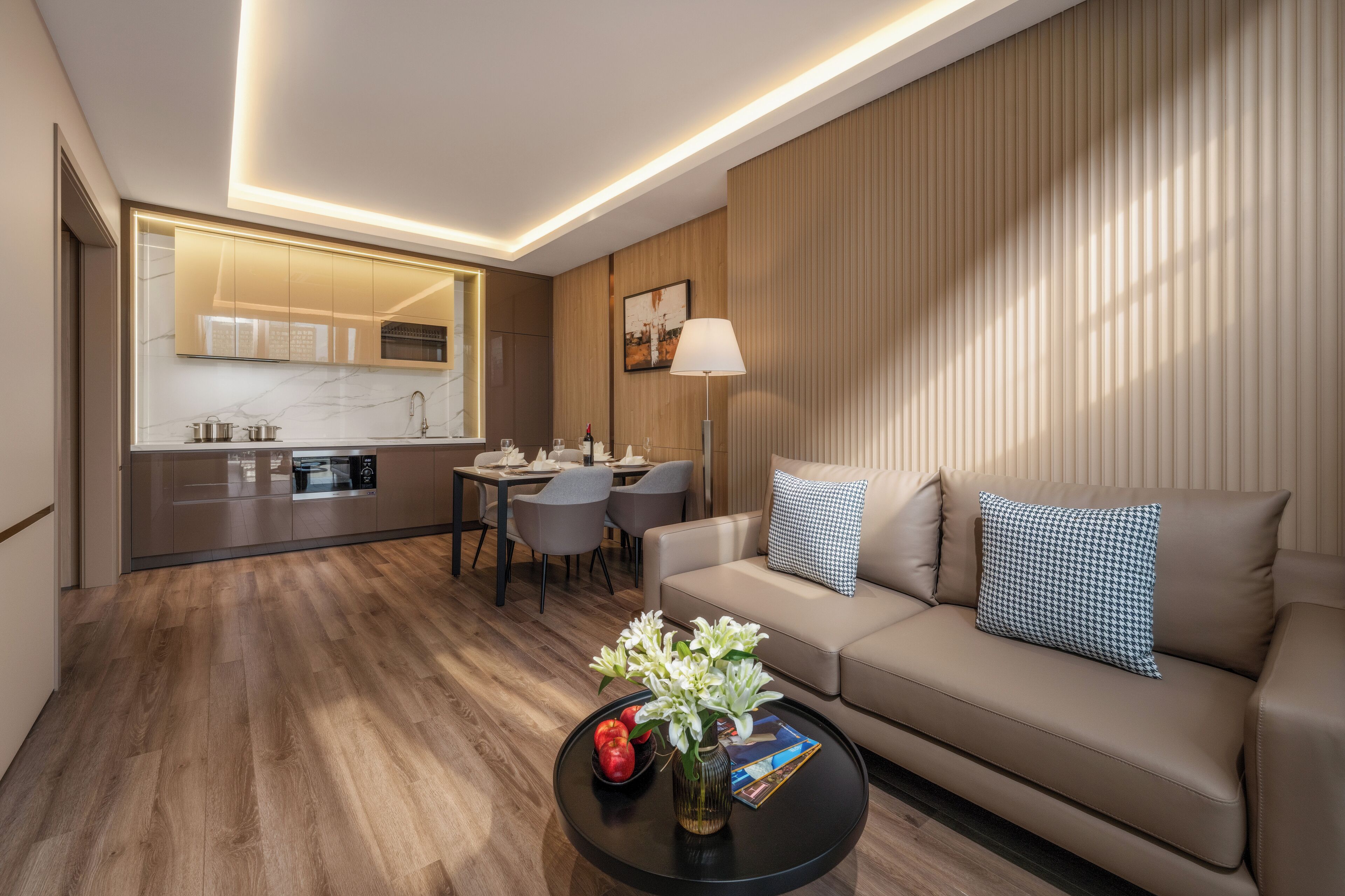 A modern hotel room with a wooden floor, a sofa, a coffee table with flowers, a dining area with chairs, and a kitchen with a mirror.