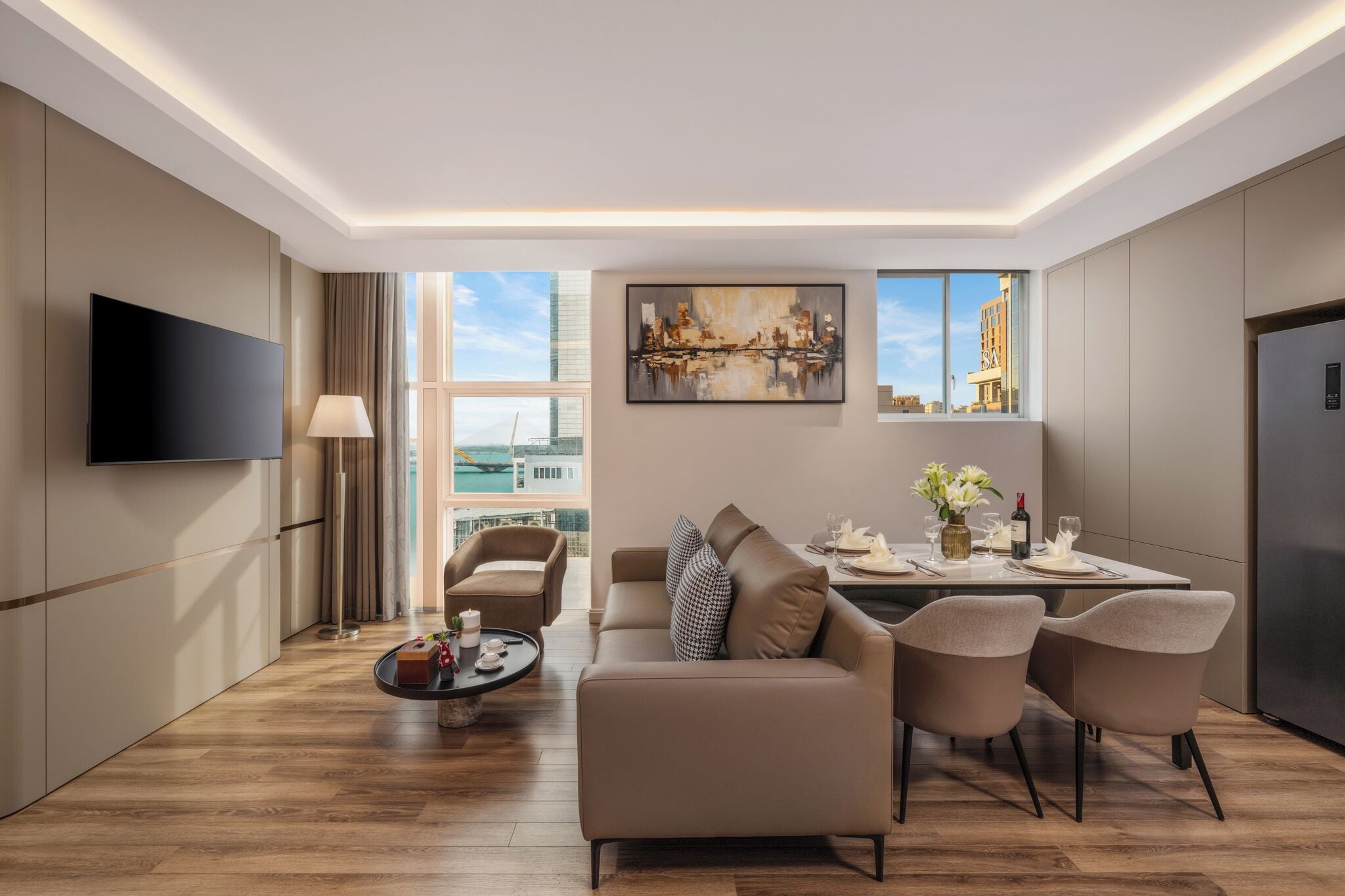 A modern hotel room with a flat-screen TV, a sofa, a dining table with chairs, and a large window offering a city view.