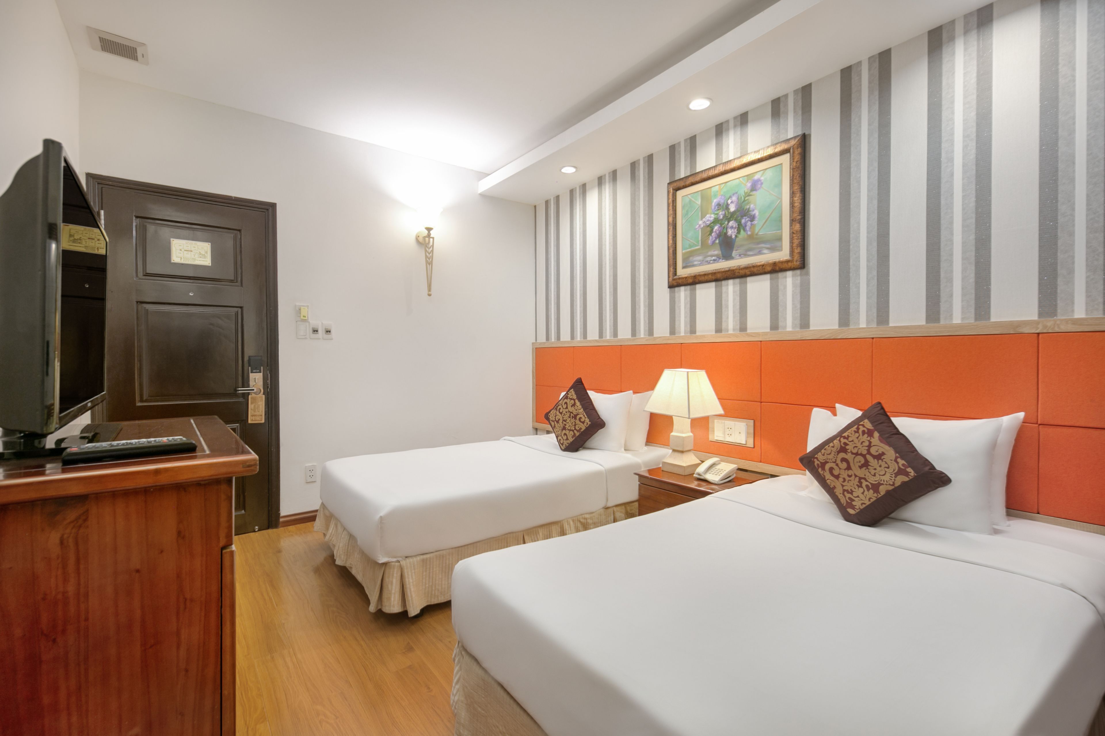 A hotel room with two single beds, a wooden desk with a television, a nightstand with a lamp, and a striped wall with a framed picture.