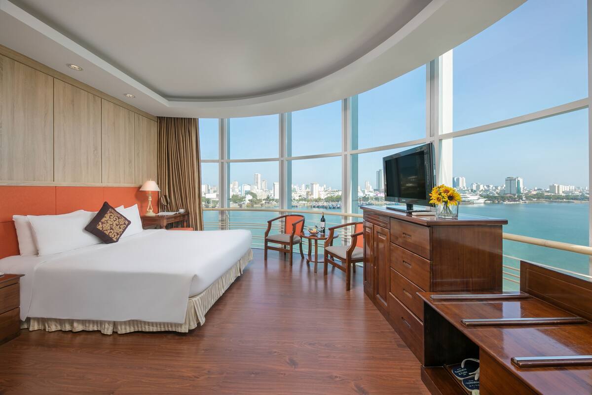 A hotel room with a large bed, a desk with a TV, a view of the city and water.
