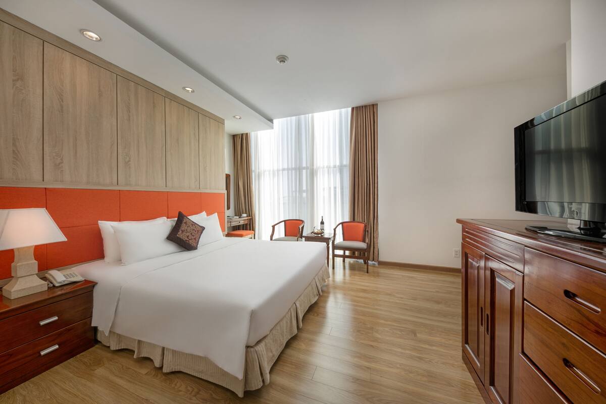 A hotel room with a large bed, wooden headboard, a TV, a desk with a chair, and a window with curtains.