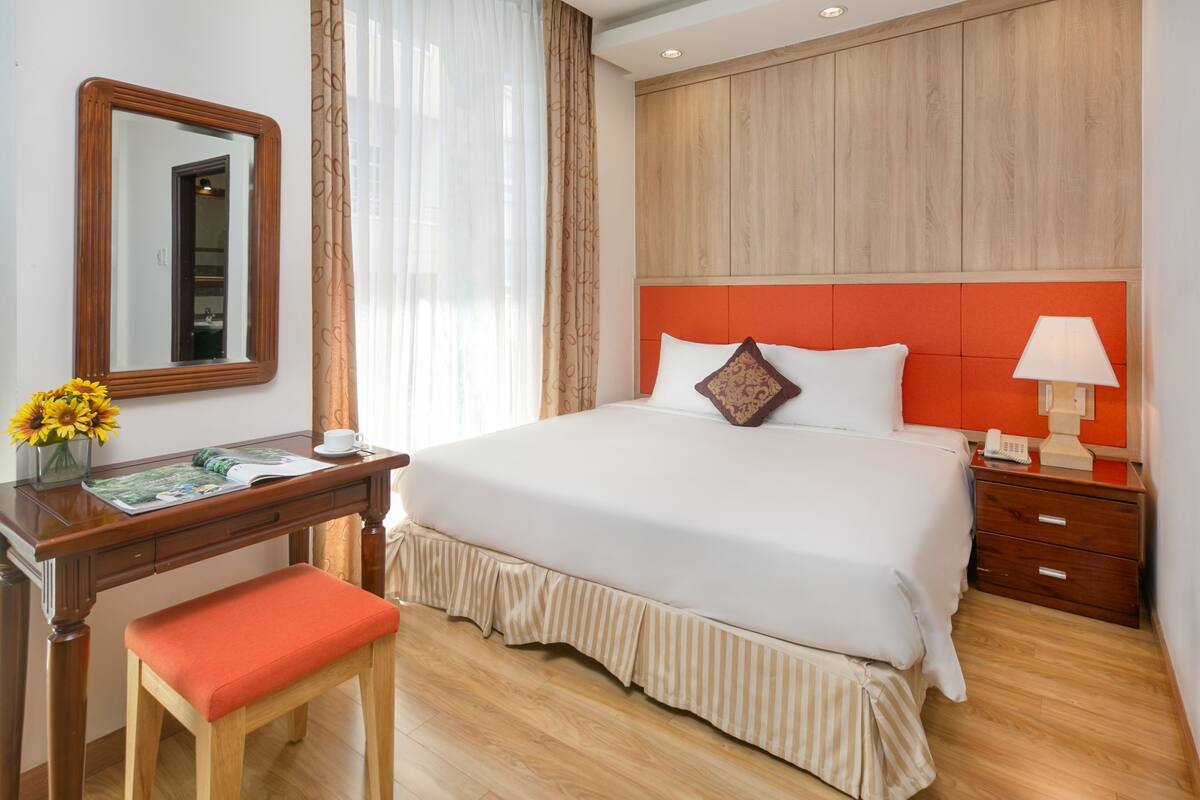 A hotel room with a large bed, a wooden desk with a chair, a bedside table with a lamp, and a mirror on the wall.