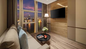 A modern hotel room with a large window offering a city view at dusk, a flat-screen TV mounted on the wall, a comfortable sofa, and a small table with a magazine and decorative items. - Sun River Hotel (Da Nang)