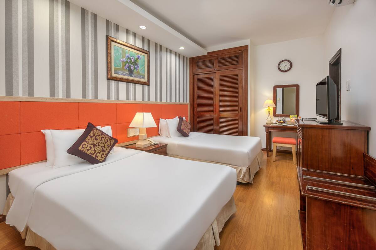 A hotel room with two beds, a wooden wardrobe, a desk with a chair, a television, and a painting on the wall.