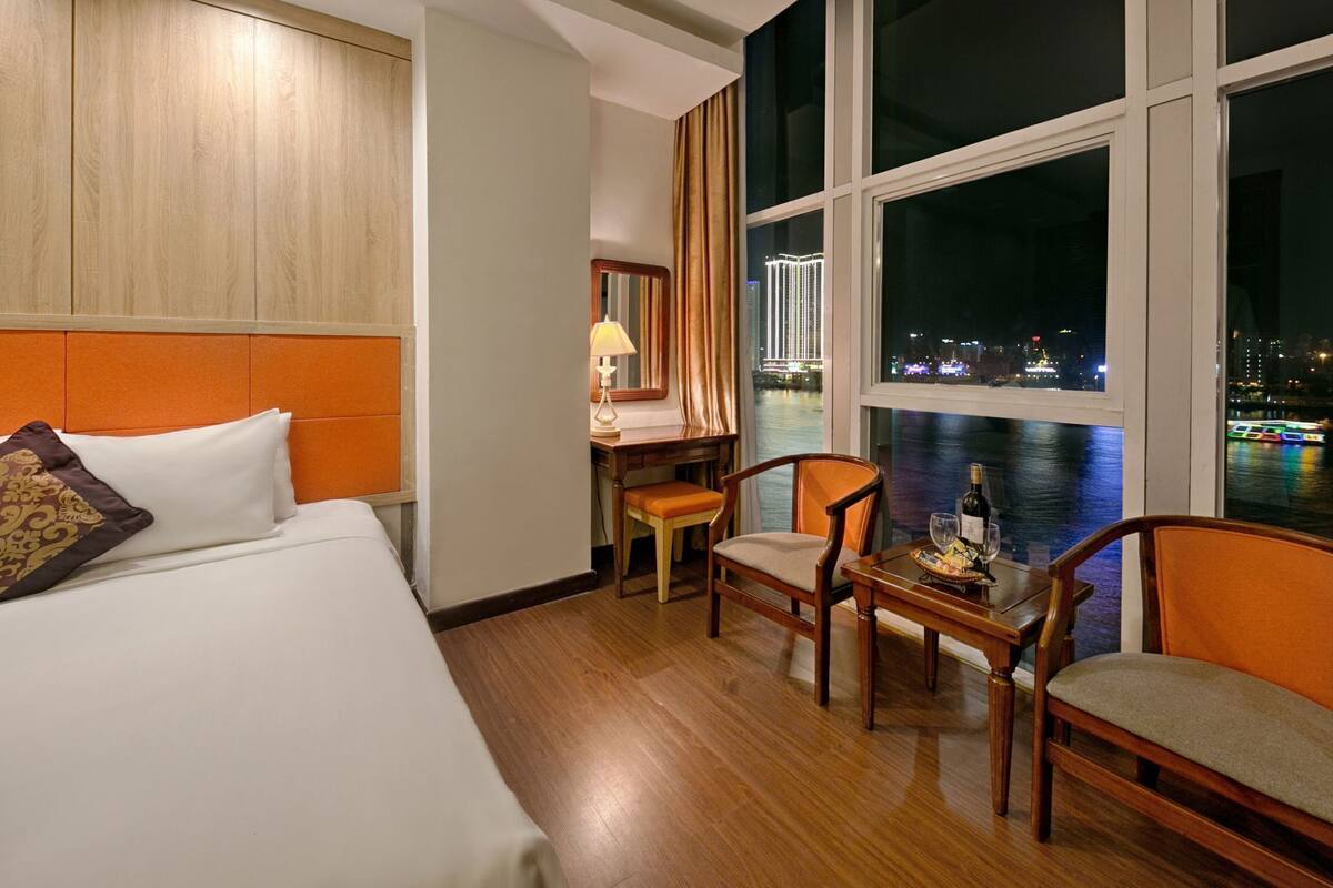 A hotel room with a bed, two chairs, a small table with a bottle and glass, and a view of a cityscape at night.