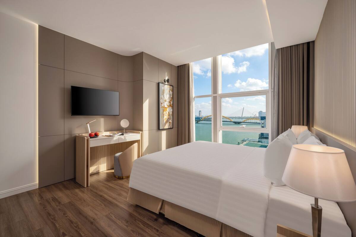 A modern hotel room with a large bed, a desk, a flat-screen TV, and a view of a bridge and water.