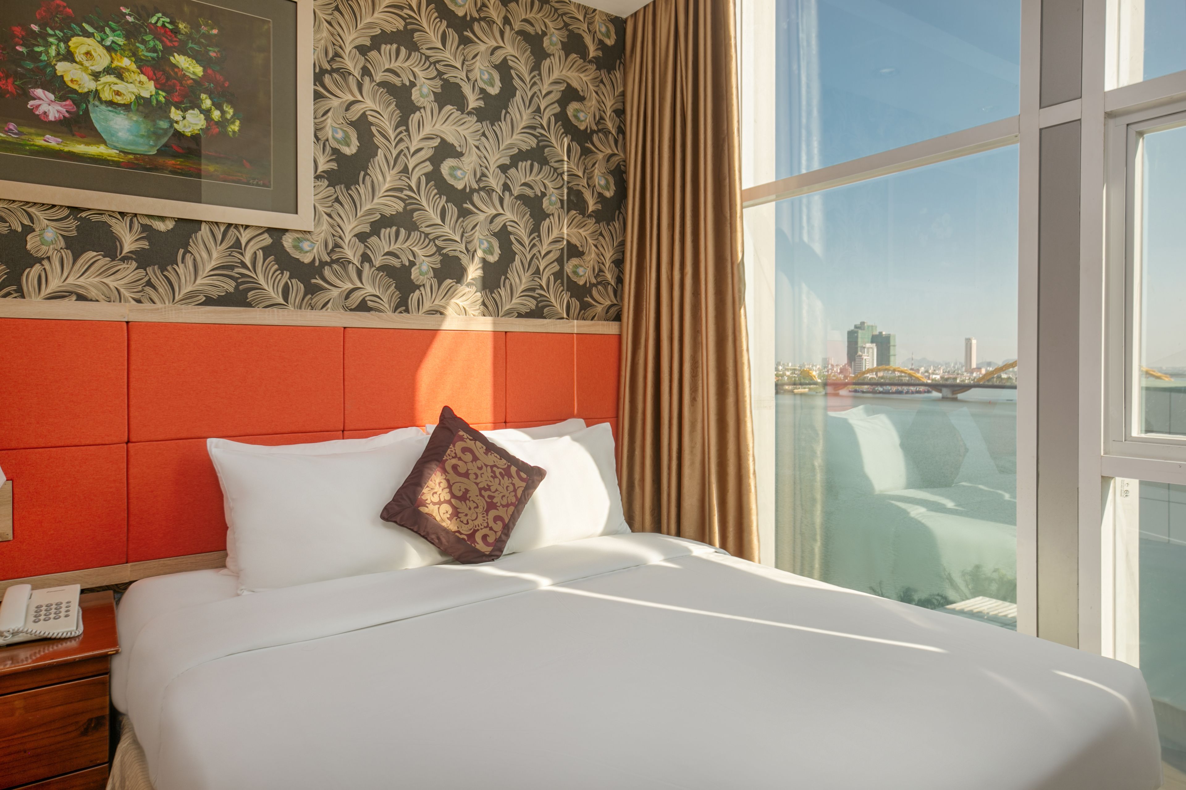 A hotel room with a large bed, a nightstand with a telephone, a window with a view of a cityscape and a bridge, and a patterned wallpaper on the wall.
