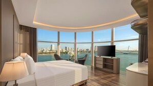 A modern hotel room with a large bed, a TV, a desk, and a view of a city skyline and water. - Sun River Hotel (Da Nang)