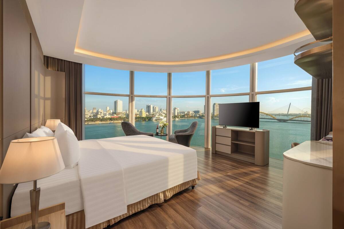 A modern hotel room with a large bed, a TV, a desk, and a view of a city skyline and water.