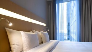 Een hotelkamer met een groot bed, twee bedlampjes en uitzicht op een gebouw door dunne gordijnen.