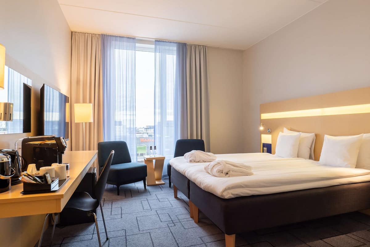 A hotel room with a bed, desk, chairs, and a view of the city through large windows.