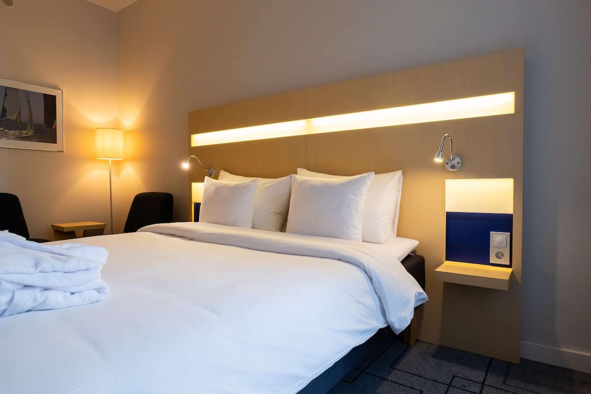 A hotel room with a large bed, a nightstand, a lamp, and a wall-mounted shelf with a mirror.