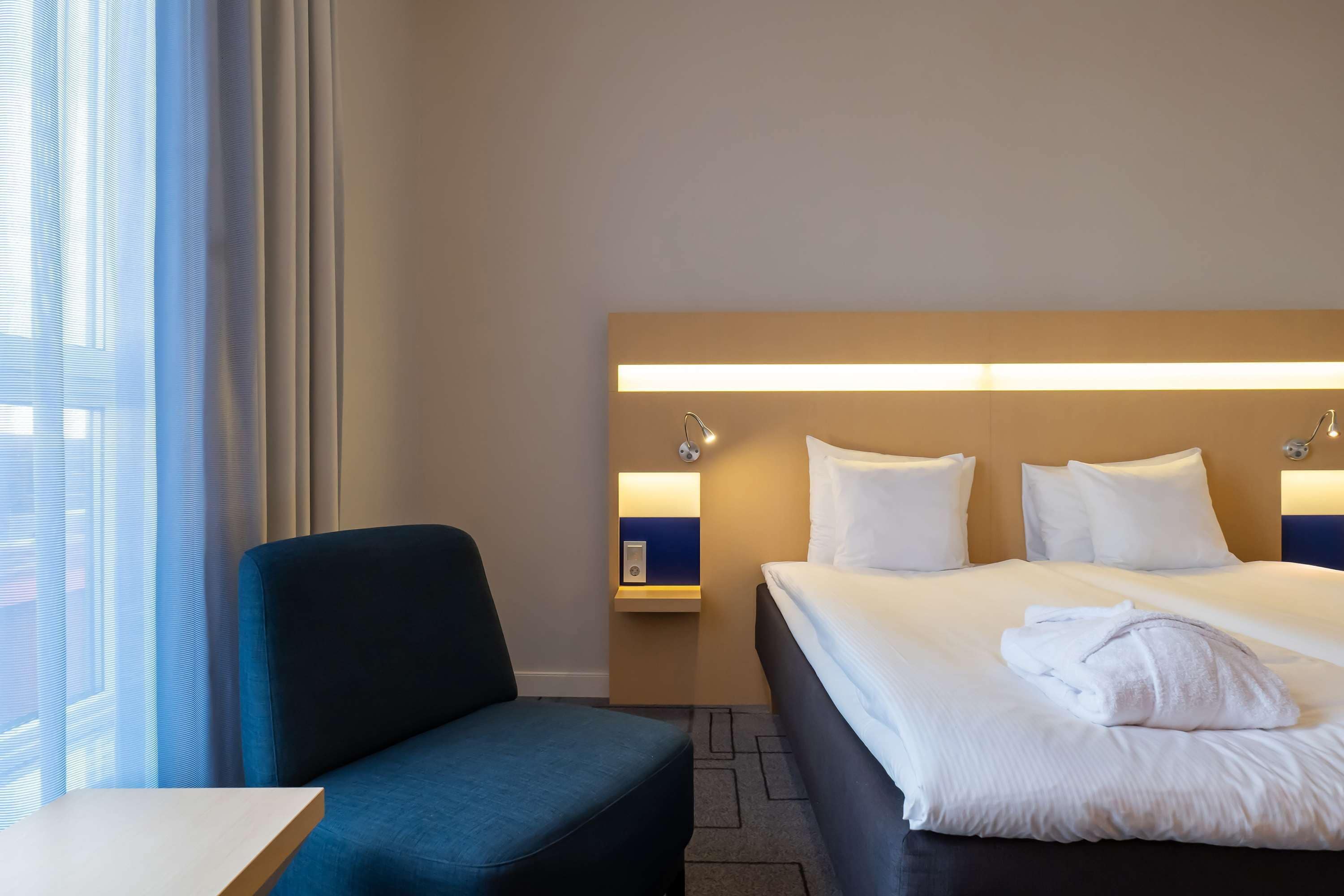 Una habitación de hotel con cama, una silla, una mesita de noche y ventana con cortinas.
