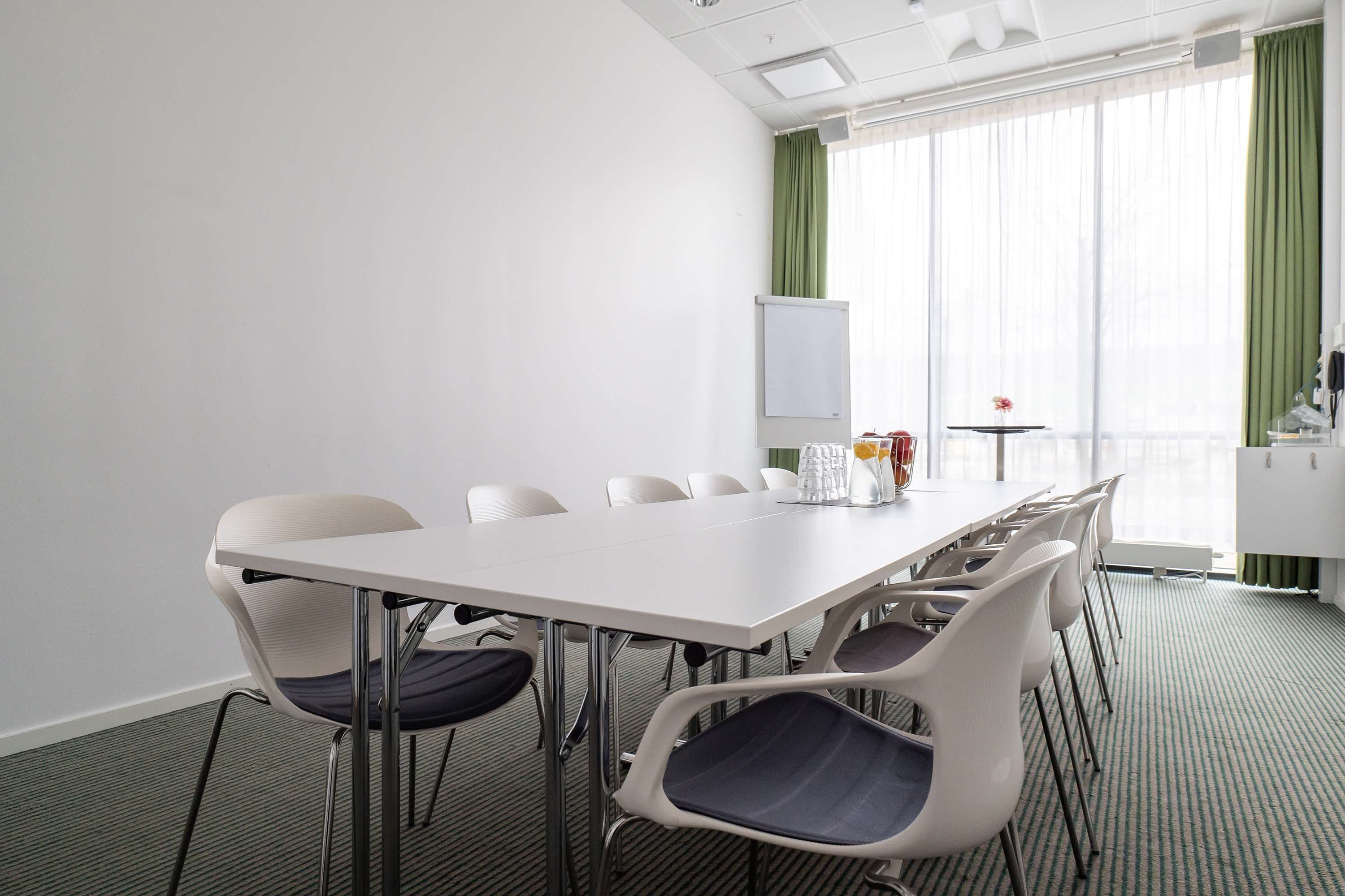 Un salón de conferencias con una mesa y sillas blancas, una pizarra y vistas a una cortina verde y una ventana.