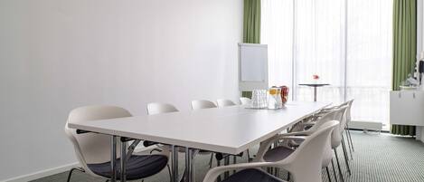Un salón de conferencias con una mesa y sillas blancas, una pizarra y vistas a una cortina verde y una ventana.