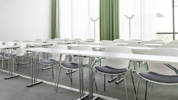 Un salón de conferencias con mesas y sillas blancas, ventanas amplias con cortinas verdes y una lámpara de pie moderna.