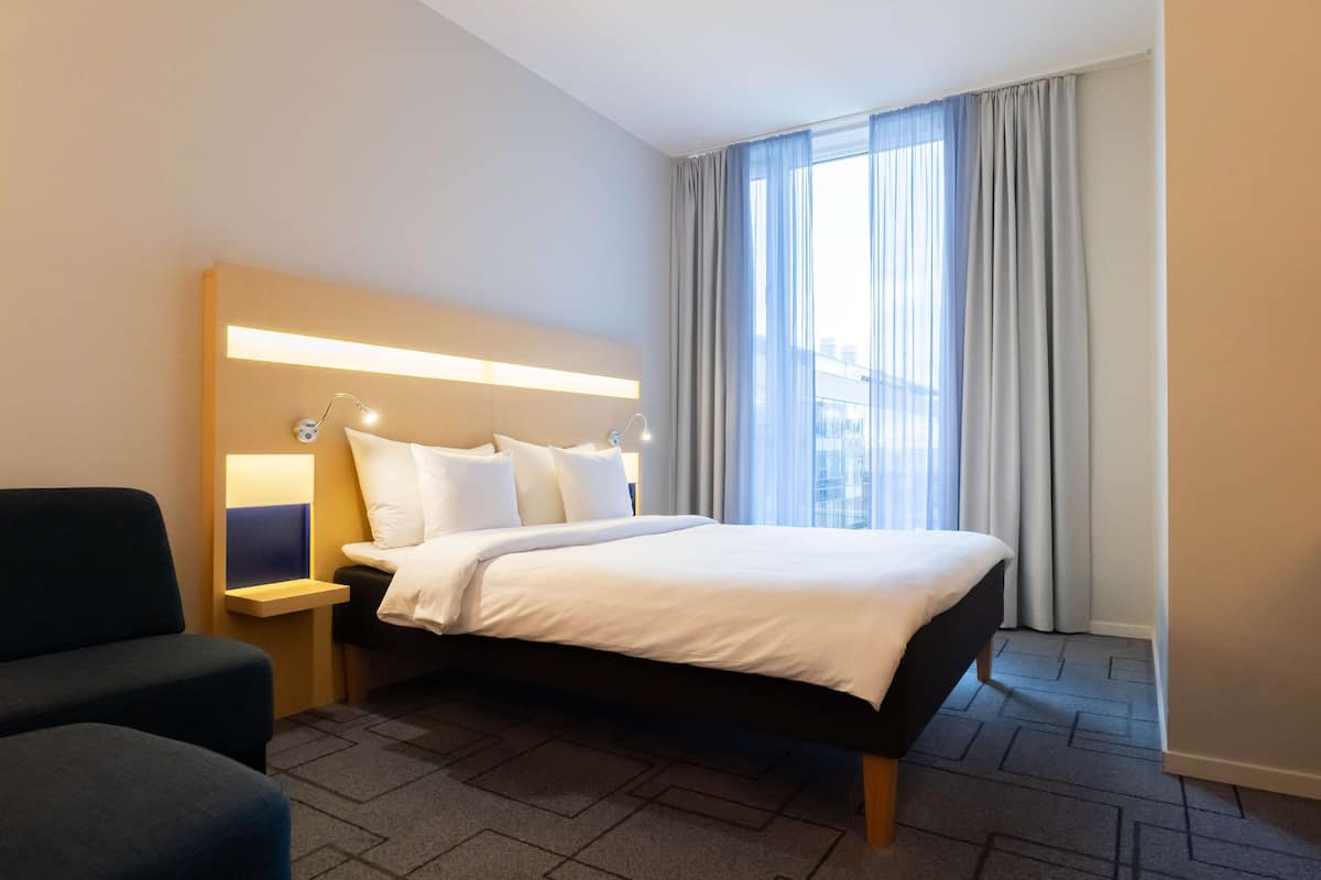A hotel room with a bed, a bedside table with a lamp, a chair, and a large window with curtains.
