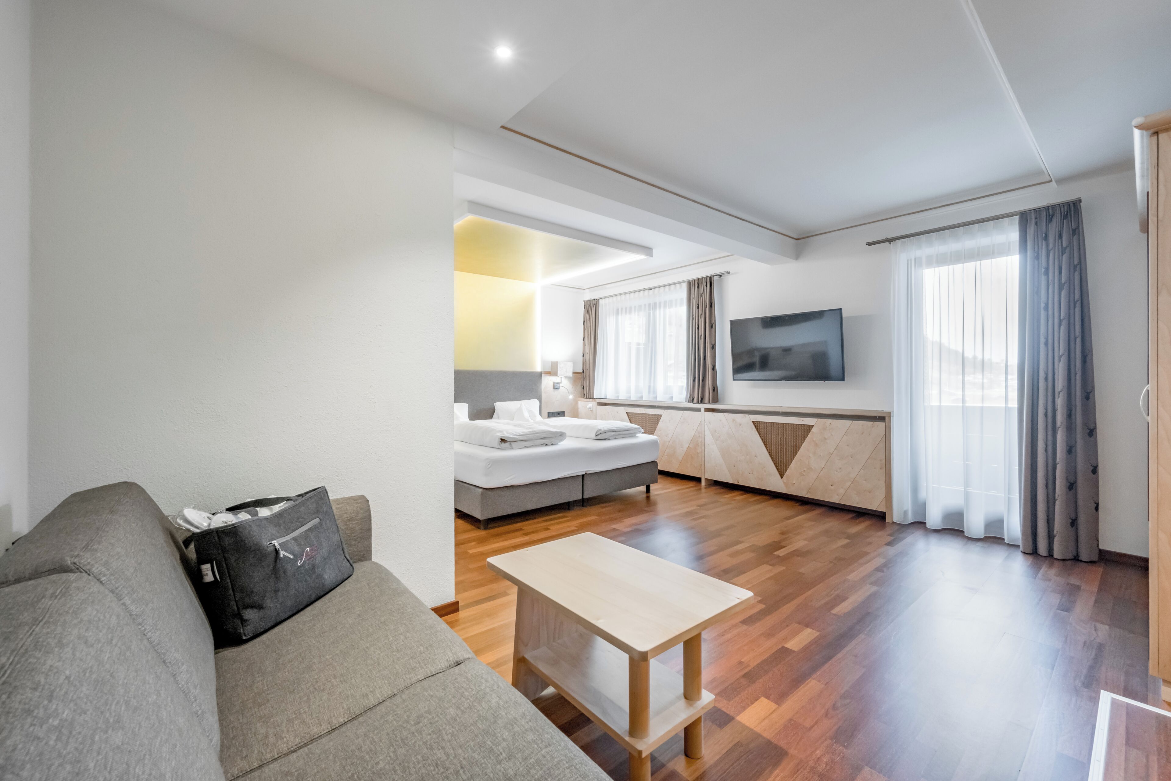 A modern living room with a sofa, a small table, a TV, and a bedroom with a bed and curtains.