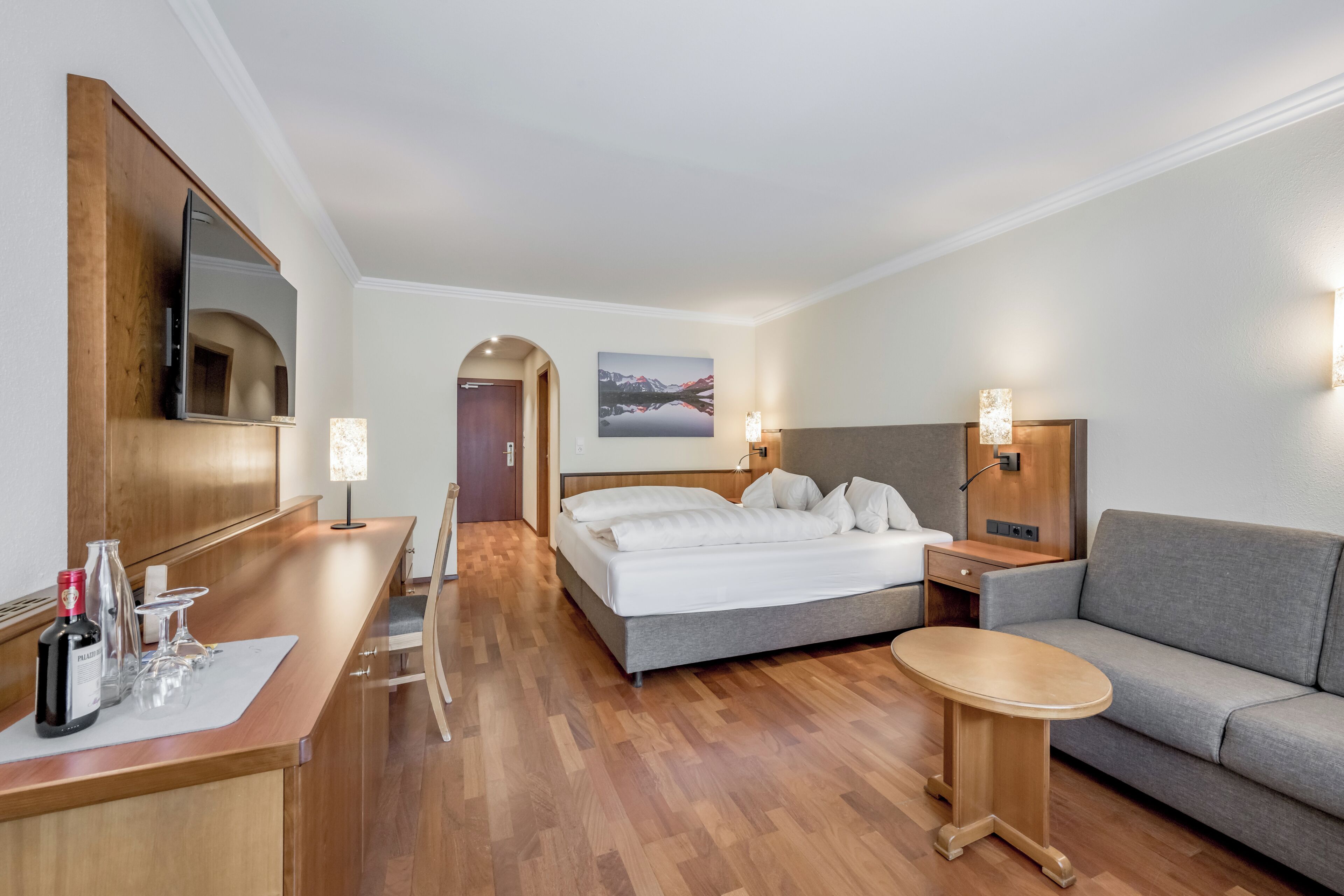 A hotel room with a bed, a sofa, a small table, a desk with a mirror, and a picture on the wall.
