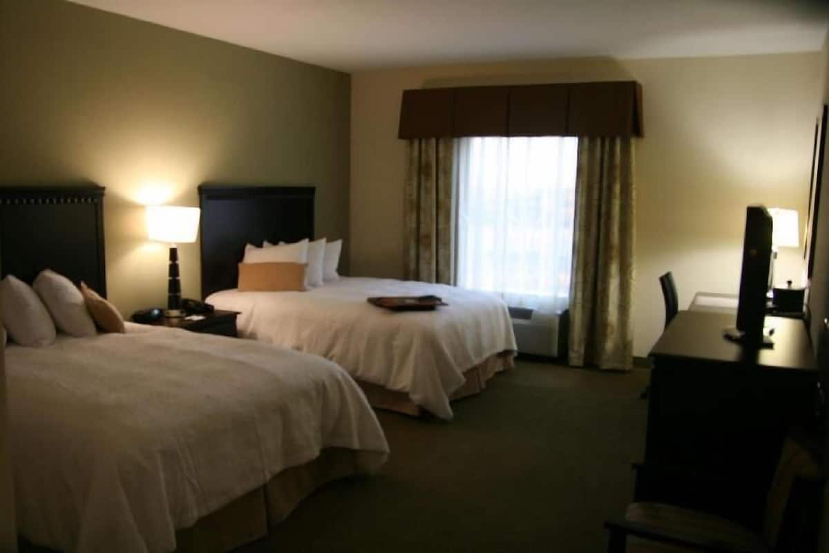 A hotel room with two beds, a desk, a chair, a lamp, and a window with curtains.