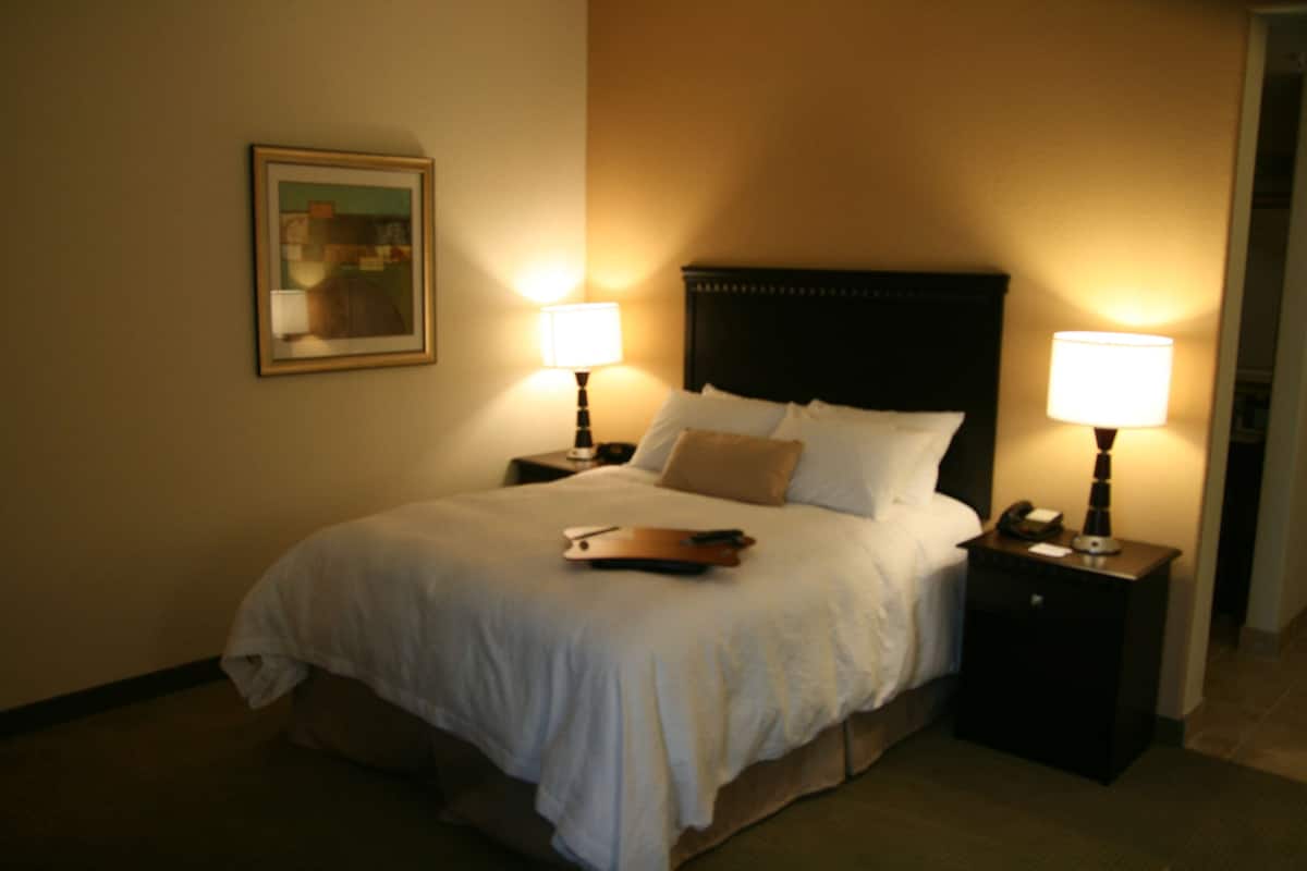 A hotel room with a bed, two bedside lamps, a nightstand, a framed picture on the wall, and a remote control on the bed.