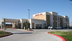 Un hotel Hampton Inn con un diseño moderno, que cuenta con un amplio estacionamiento y un cielo despejado.