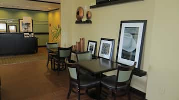 Um saguão de hotel com uma mesa de recepção, uma área de refeições com uma mesa e cadeiras, e uma televisão na parede.
