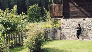 A rustic wooden building with a stone wall, surrounded by a wooden fence and greenery, set against a backdrop of a forested mountain. - Posthotel (Valle Aurina)