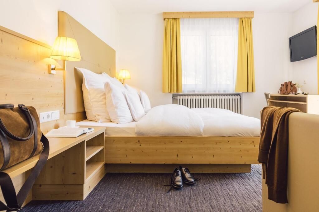 A hotel room with a bed, bedside table, lamp, window with curtains, radiator, and a TV on the wall.