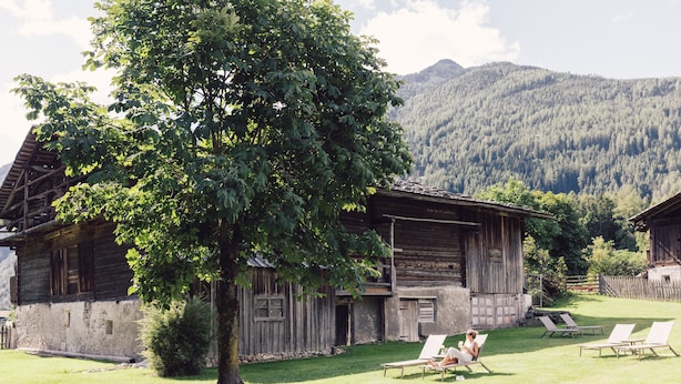 A rustic wooden house with a large tree in front, a grassy yard, and a mountain in the background. - Posthotel (Valle Aurina)