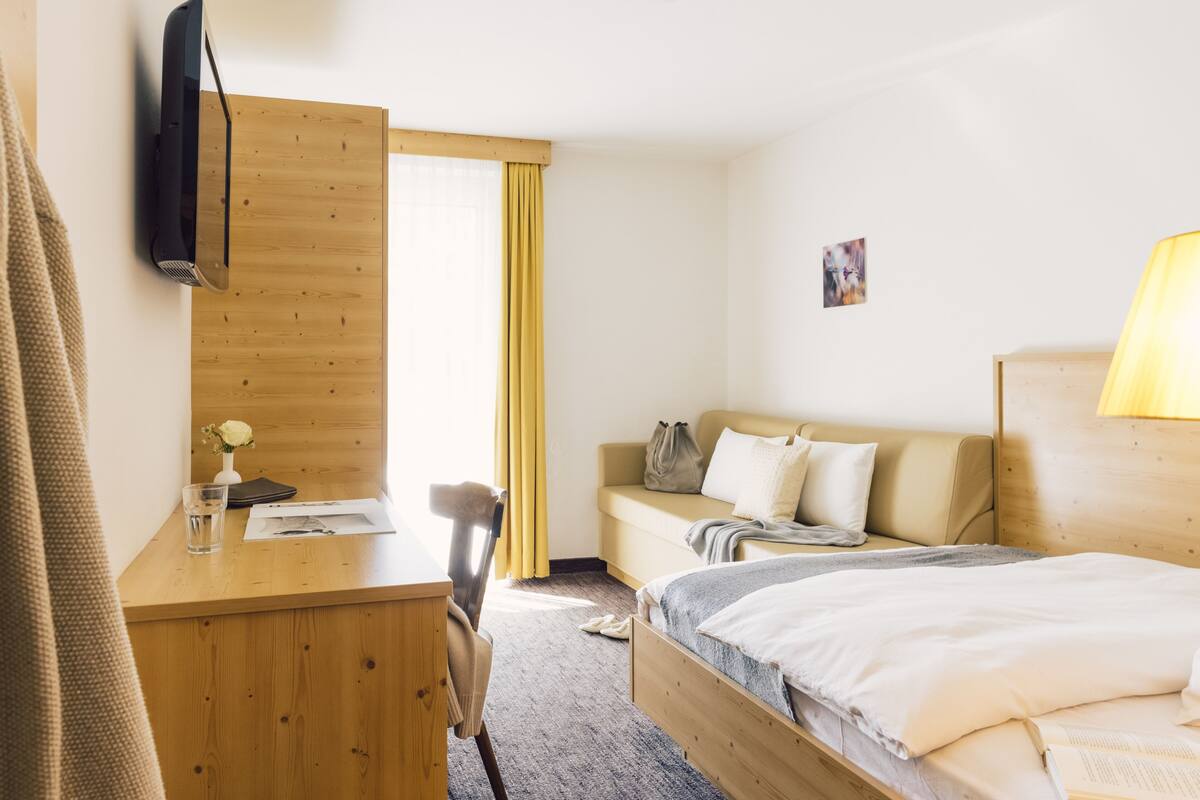 A hotel room with a wooden desk, a bed with white bedding, a sofa, and a TV mounted on the wall.