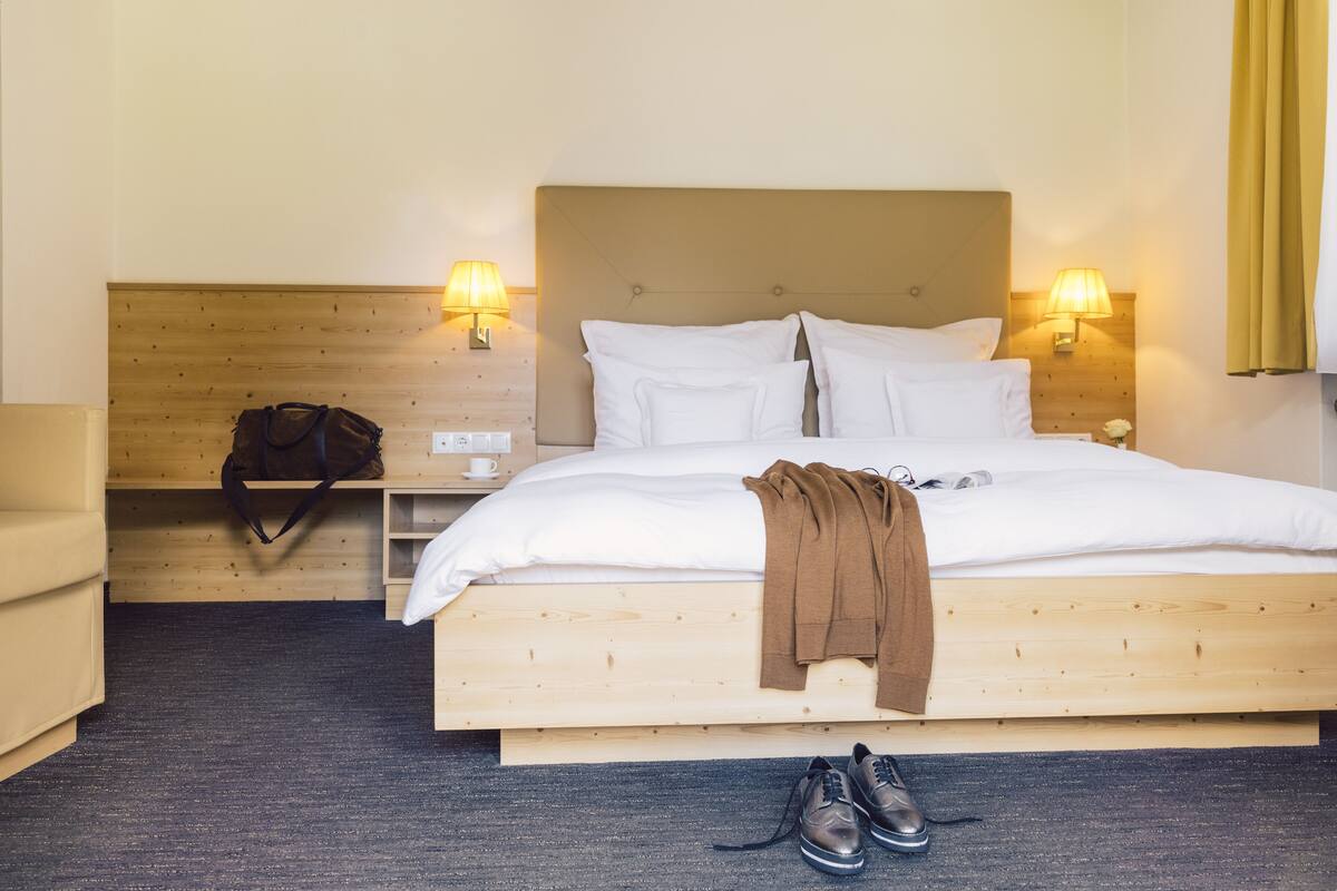 A neatly made bed with white linens, a brown throw blanket, and a pair of shoes on the floor. There are two bedside lamps, a wooden headboard, and a small table with a cup and a bag.