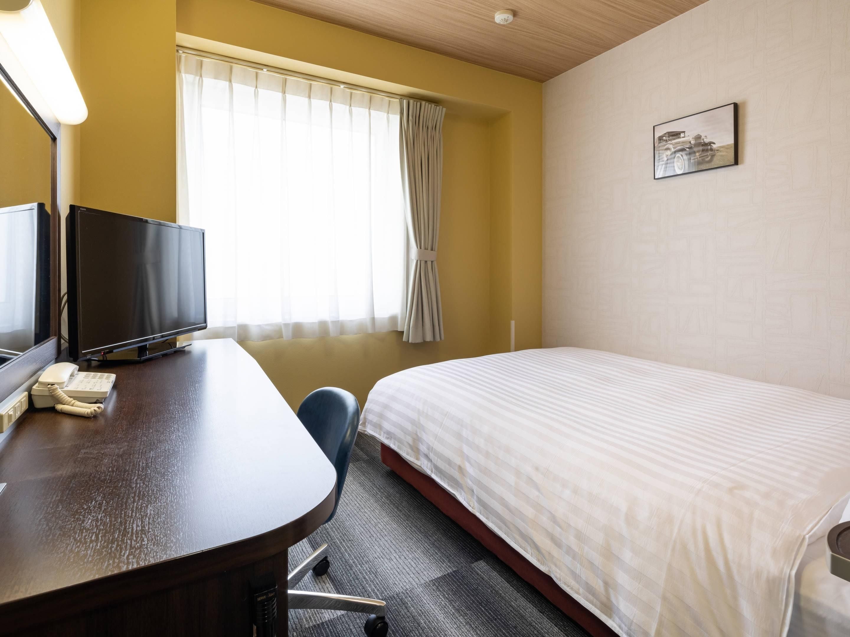 A hotel room with a bed, a desk with a television and phone, a chair, and a window with curtains.