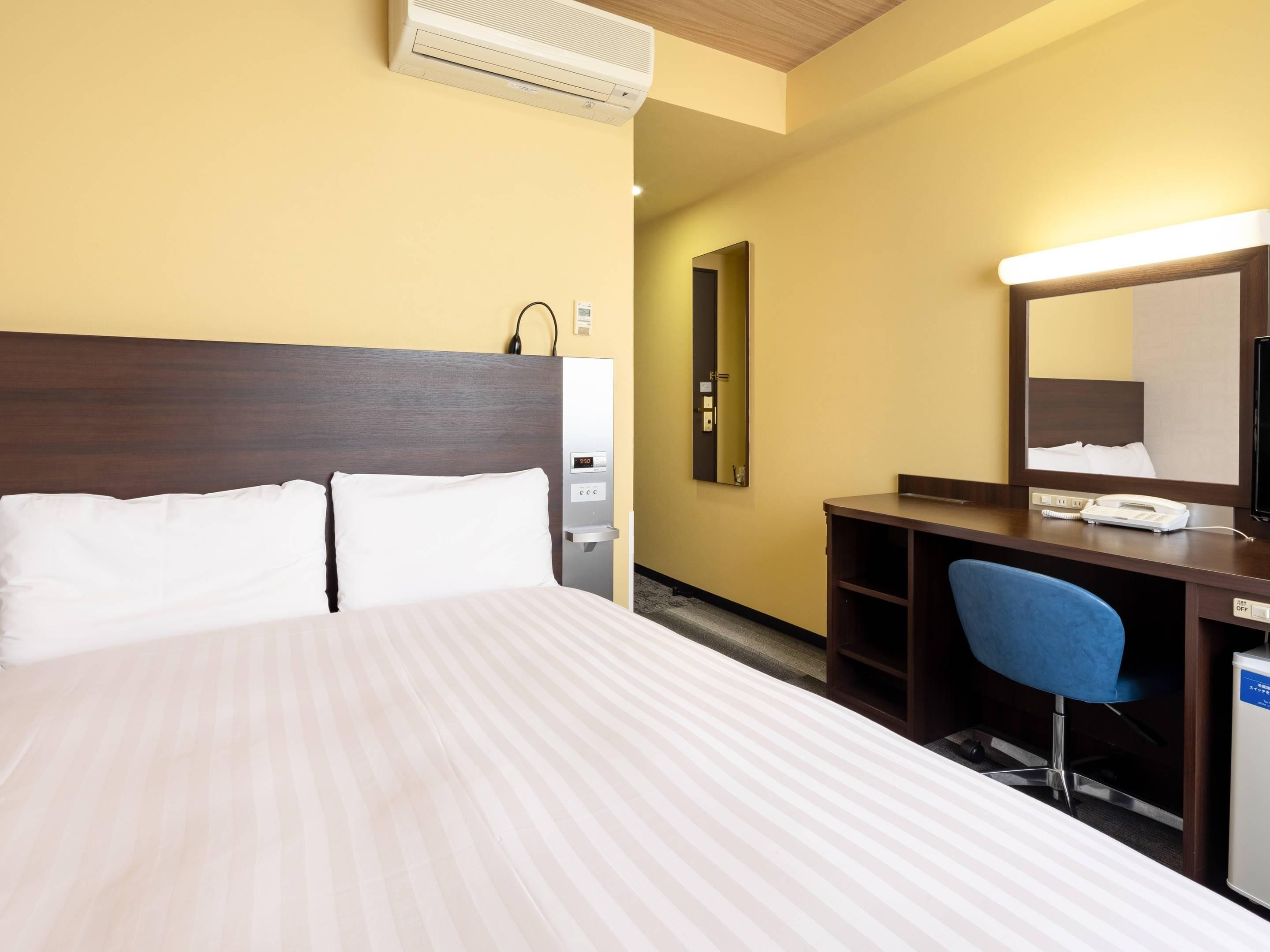 Habitación de hotel con cama, escritorio, silla, espejo y aire acondicionado instalado en la pared.