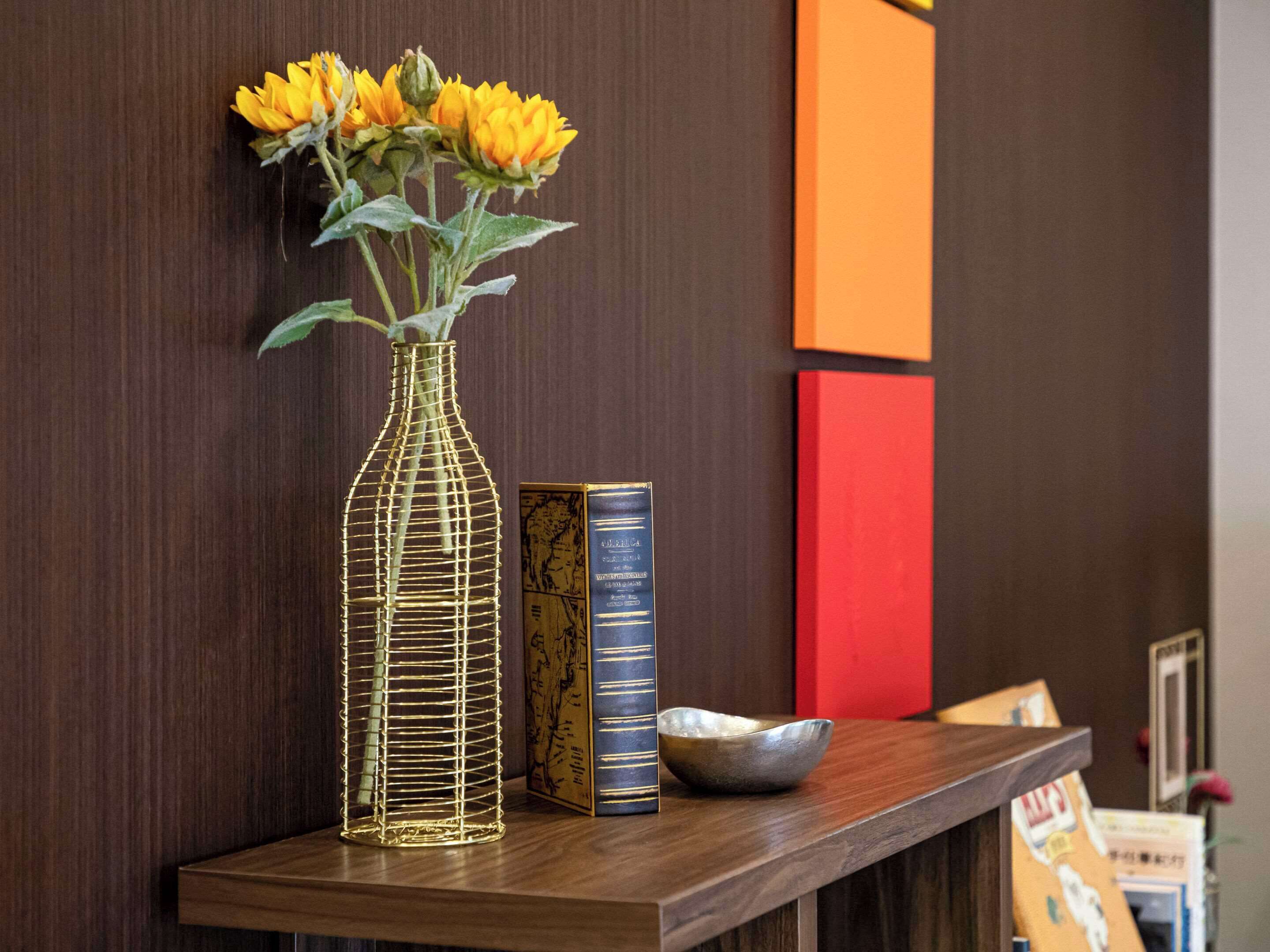 A wooden shelf with a wireframe vase containing yellow flowers, a book, and a ceramic bowl.