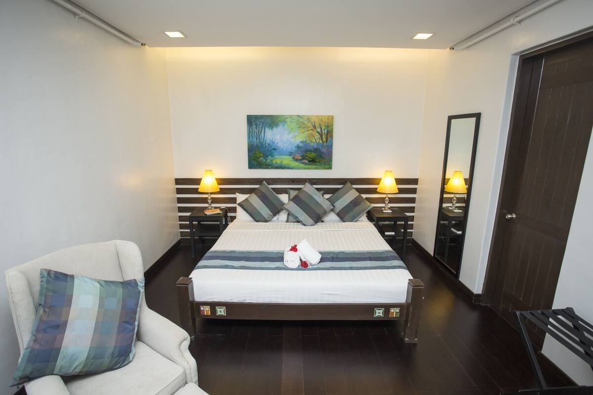 Un cuarto de hotel con una cama grande, dos mesitas de noche con lámparas, una silla, un cuadro en la pared y una puerta.