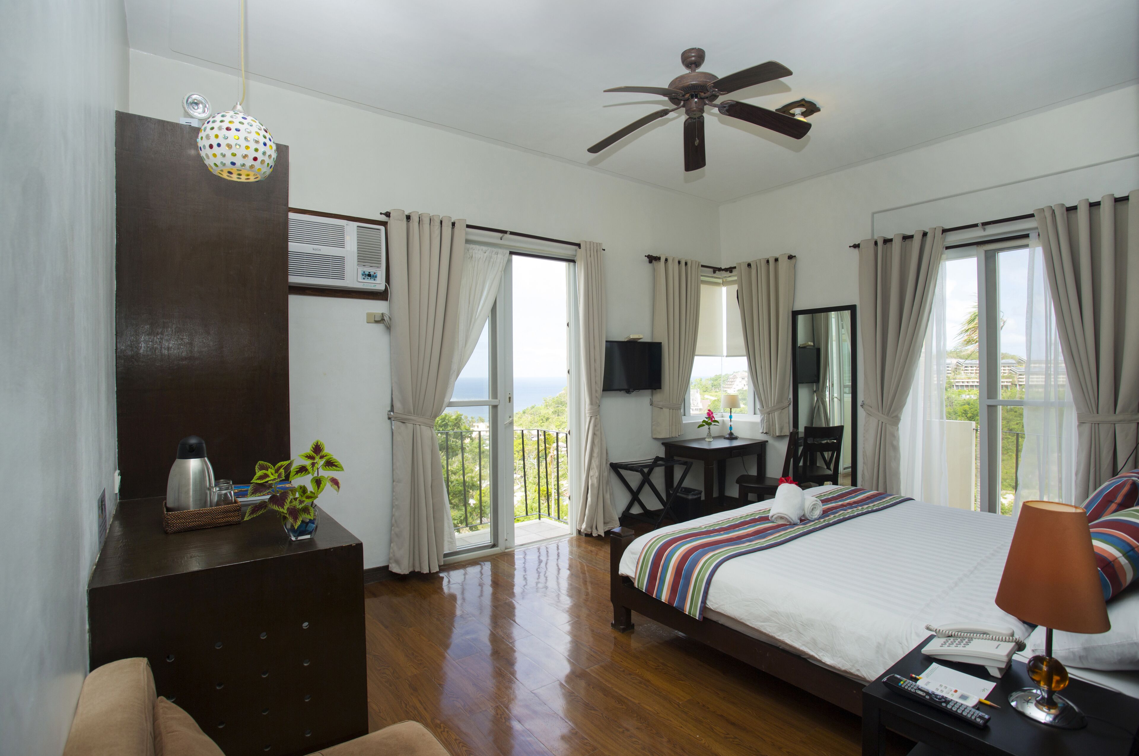 Habitación de hotel con cama, escritorio, silla, televisor y balcón con cortinas.