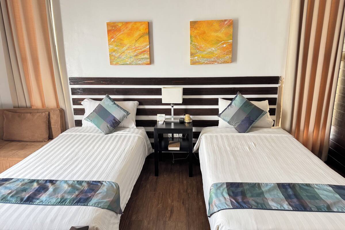 Habitación de hotel con dos camas, una mesita de noche con lámpara y dos cuadros abstractos en la pared.