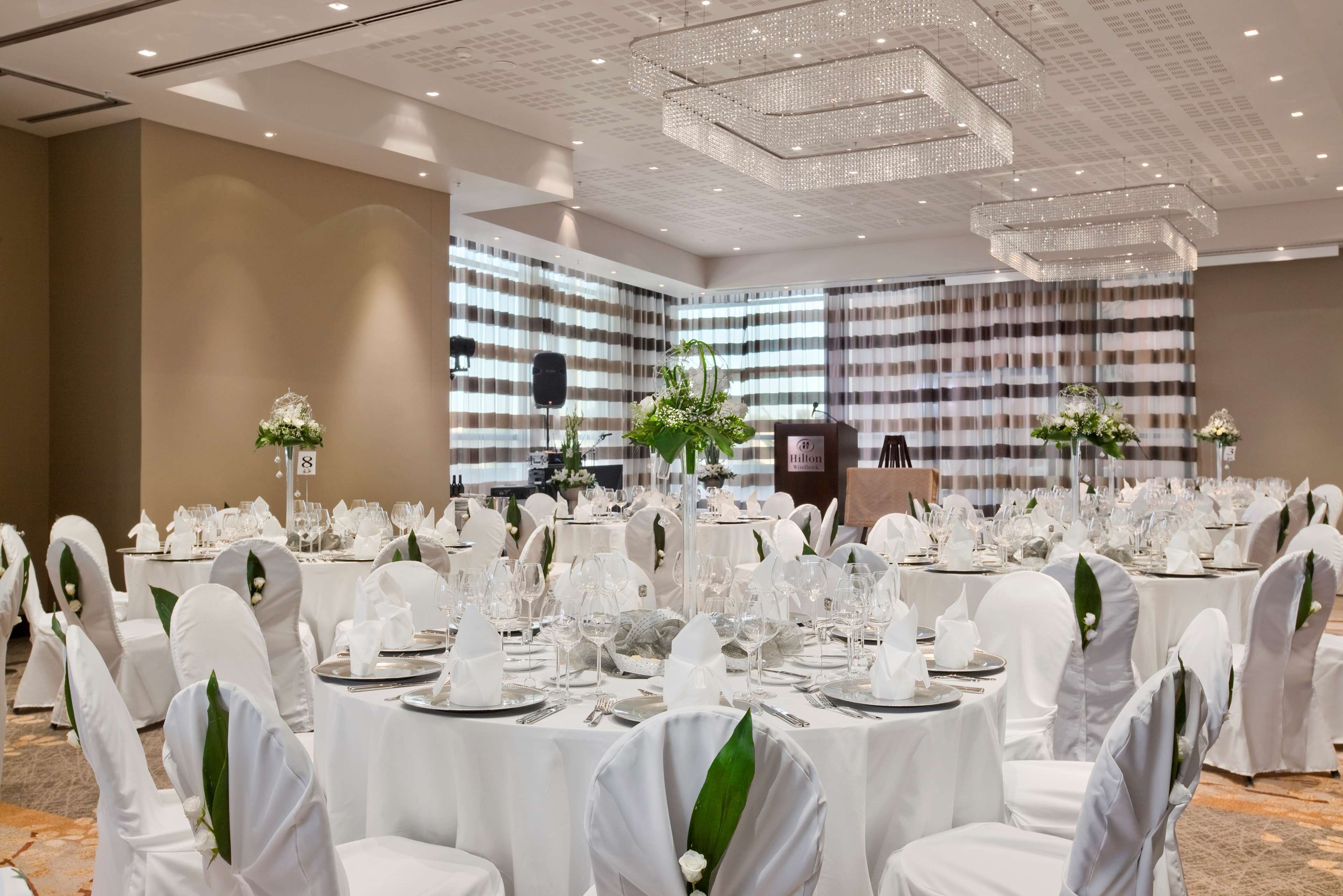 A banquet hall with round tables set for a formal event, featuring white tablecloths, chairs with white covers, and floral centerpieces.