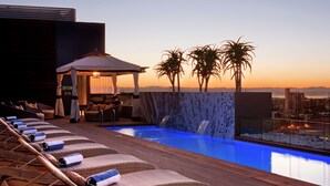A rooftop pool area with lounge chairs, a waterfall, and a view of the city at sunset. - Hilton Windhoek (Windhoek)