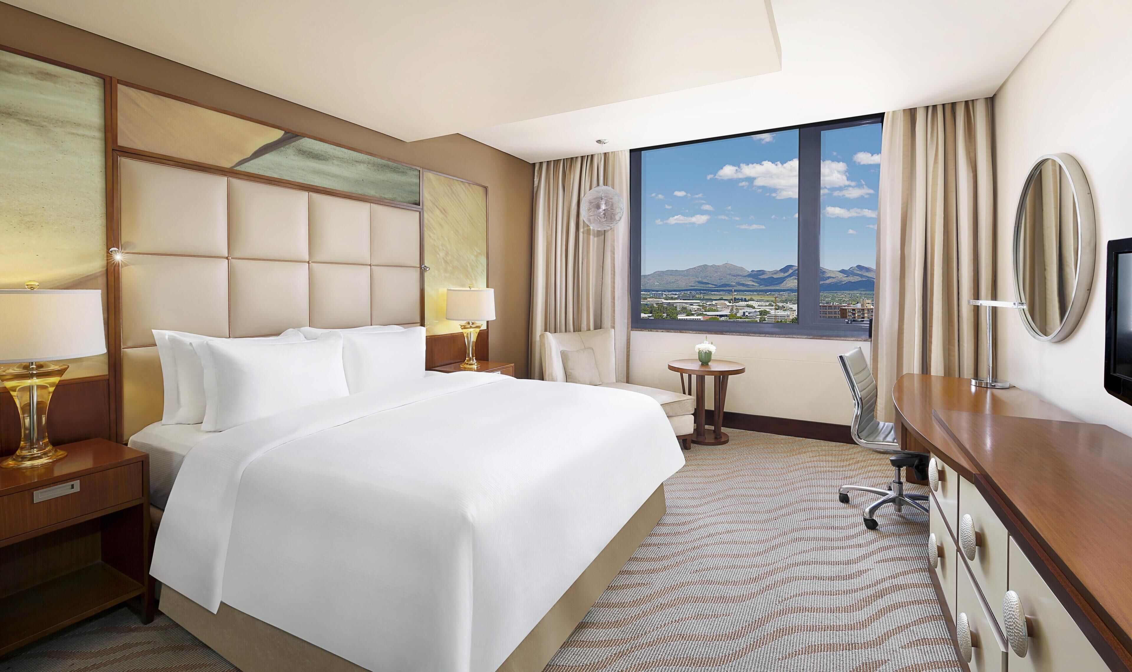 A hotel room with a large bed, a desk, a chair, a small table, a lamp, a mirror, and a view of the city and mountains through the window.